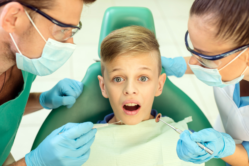 Bambino su sedia da dentista, con dentista e assistente ai suoi fianchi; concept: carie