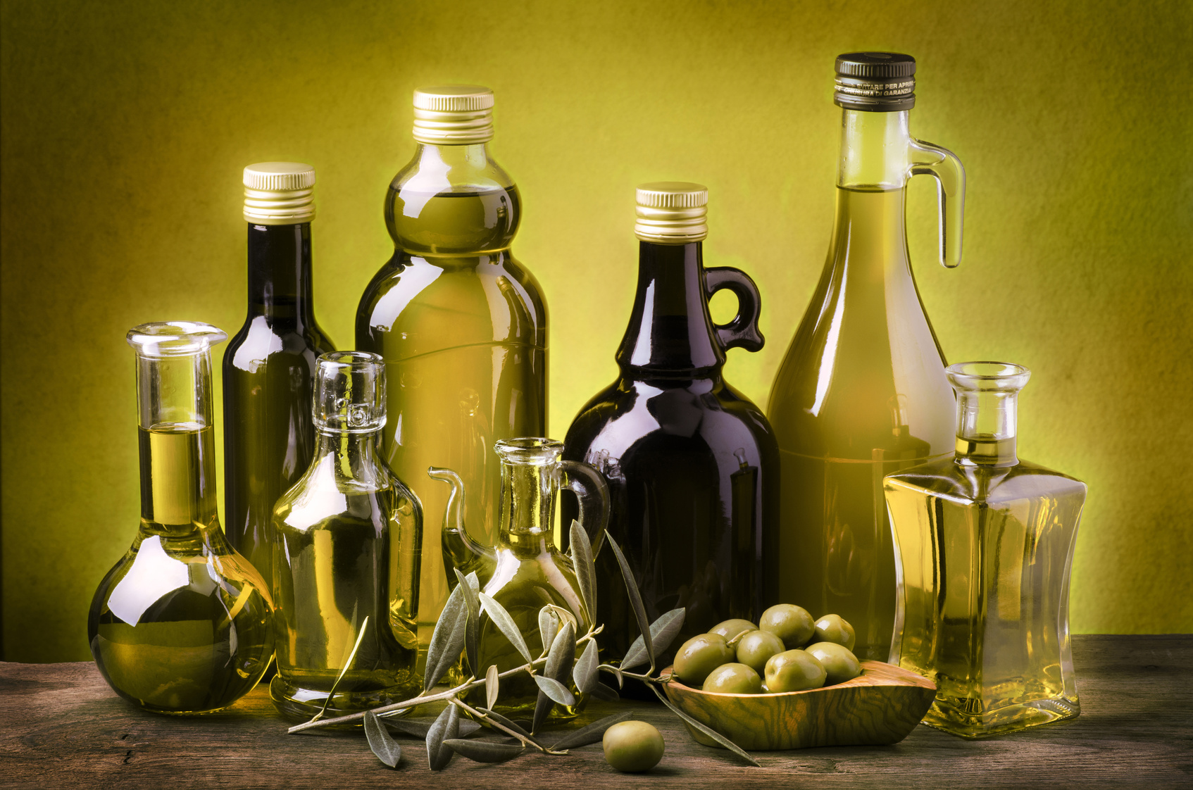 Bottiglie di olio extravergine oliva di varie forme e colori, con olive e ramo di ulivo