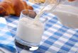 latte, caraffa di vetro che versa latte in bicchiere di vetro su tovaglia a quadretti bianca e blu, sullo sfondo si vede un croissant