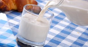 latte, caraffa di vetro che versa latte in bicchiere di vetro su tovaglia a quadretti bianca e blu, sullo sfondo si vede un croissant