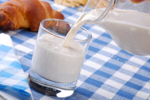 latte, caraffa di vetro che versa latte in bicchiere di vetro su tovaglia a quadretti bianca e blu, sullo sfondo si vede un croissant