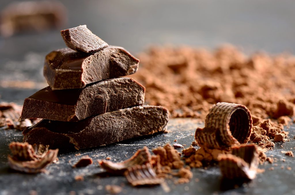 Cioccolato fondente a pezzi su un tavolo in mezzo a riccioli di cioccolato e cacao in polvere