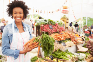 Donna con grembiule mostra mazzo di carote accanto a banco di frutta e verdura del mercato