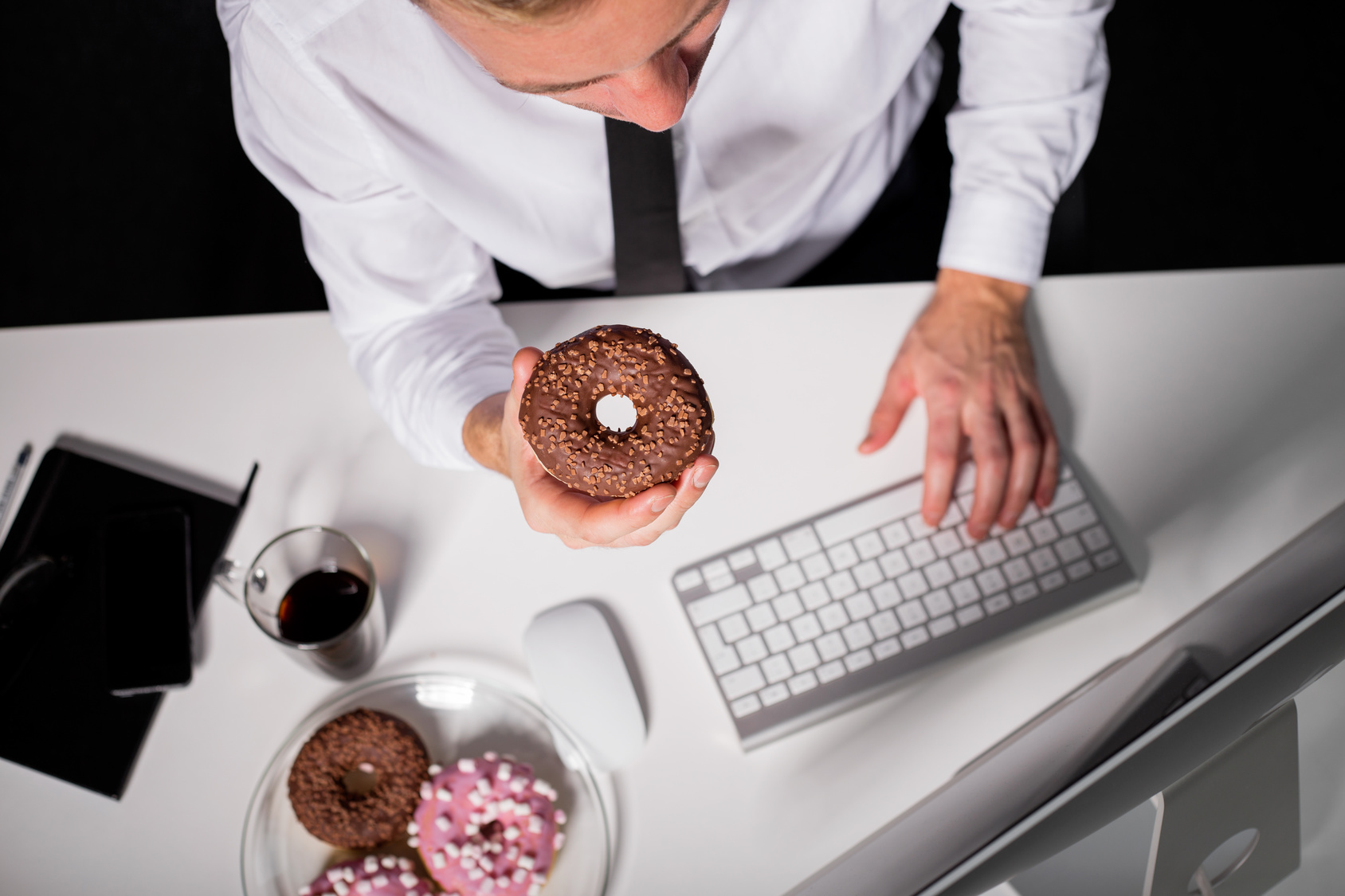 Uomo mangia ciambelle o donut glassate alla scrivania davanti al computer: concept: junk food, lavoro