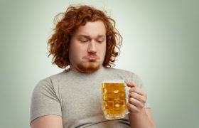 Uomo in sovrappeso guarda con espressione imbronciata un boccale di birra; concept: alcol, alcolici