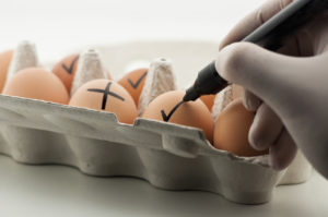 Persona con mano guantata segna uova in un cartone con un pennarello con X o segni di spunta; concept: contaminazione, salmonella, allerta
