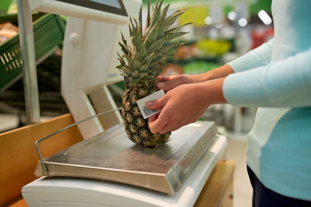 Donna etichetta un ananas dopo averlo pesato sulla bilancia del supermercato; concept: ortofrutta, frutta esotica