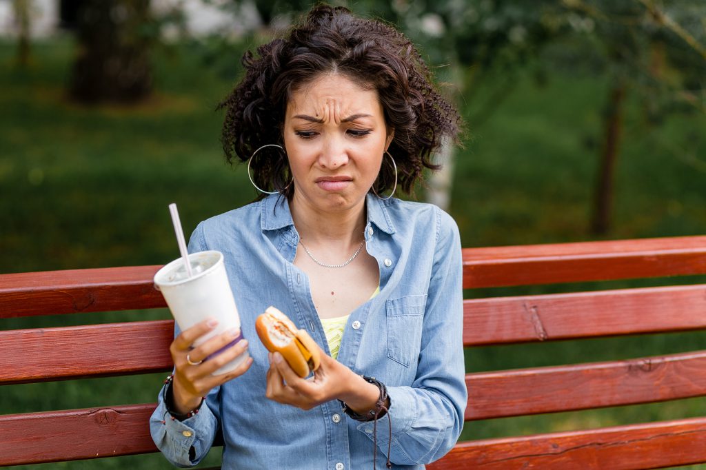 Outdoor portrait of woman looking at fast food hamburger and soda with disgusting emotion