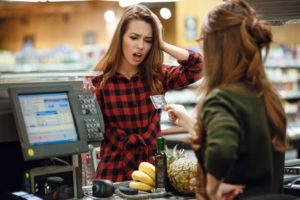 Cliente alla cassa del supermercato esprime shock mentre la cassiera porge carta di credito; prodotti sul nastro trasportatore