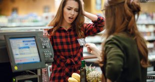 Cliente alla cassa del supermercato esprime shock mentre la cassiera porge carta di credito; prodotti sul nastro trasportatore