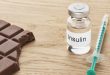 Eine Insulinampulle steht zusammen mit einer Spritze auf einem Tisch daneben liegt ein angebissenes stück Schokolade.