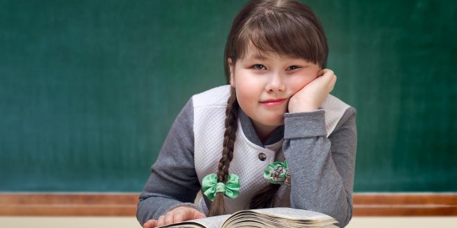 Bambina sovrappeso appoggiata su un libro sulla cattedra, davanti alla lavagna