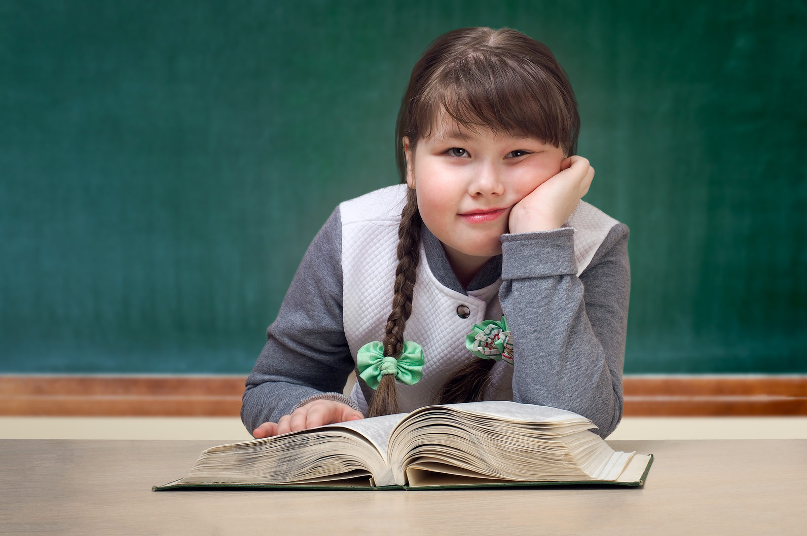 Bambina sovrappeso appoggiata su un libro sulla cattedra, davanti alla lavagna