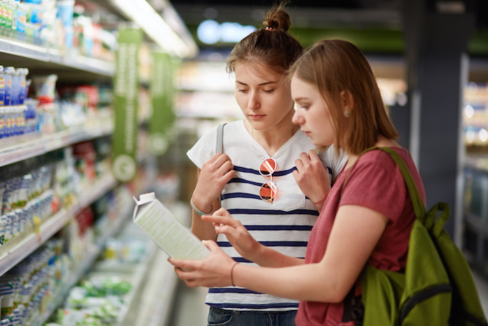 Two pretty female sisters go shopping together, stands in grocer`s shop, select fresh milk in paper container, read label, carry rucksacks, have serious expressions. People and commerce concept supermercato etichetta allergeni