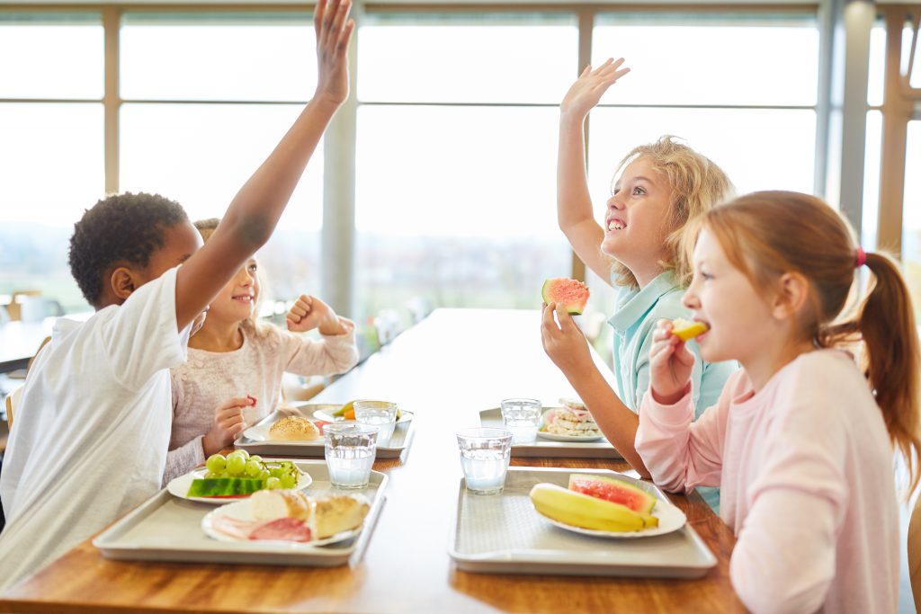Bambini al tavolo di una mensa scolastica con vassoi di frutta, verdura, pane e carne o salumi