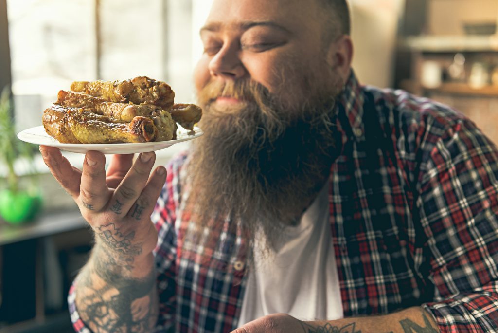 Uomo con camicia a quadri e barba lunga annusa soddisfatto piatto di cosce di pollo