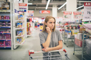 Donna con espressione dubbiosa appoggiata a un carrello tra le corsie di un supermercato