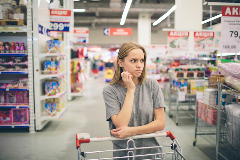 Donna con espressione dubbiosa appoggiata a un carrello tra le corsie di un supermercato