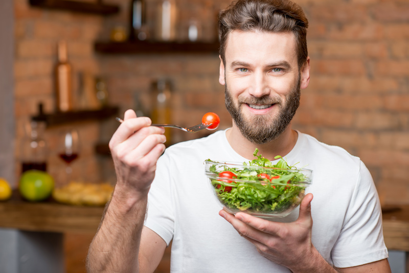 Uomo sorridente tiene in una mano ciotola di vetro con insalata verde e pomodorini e nell'altra una forchetta con un pomodorino