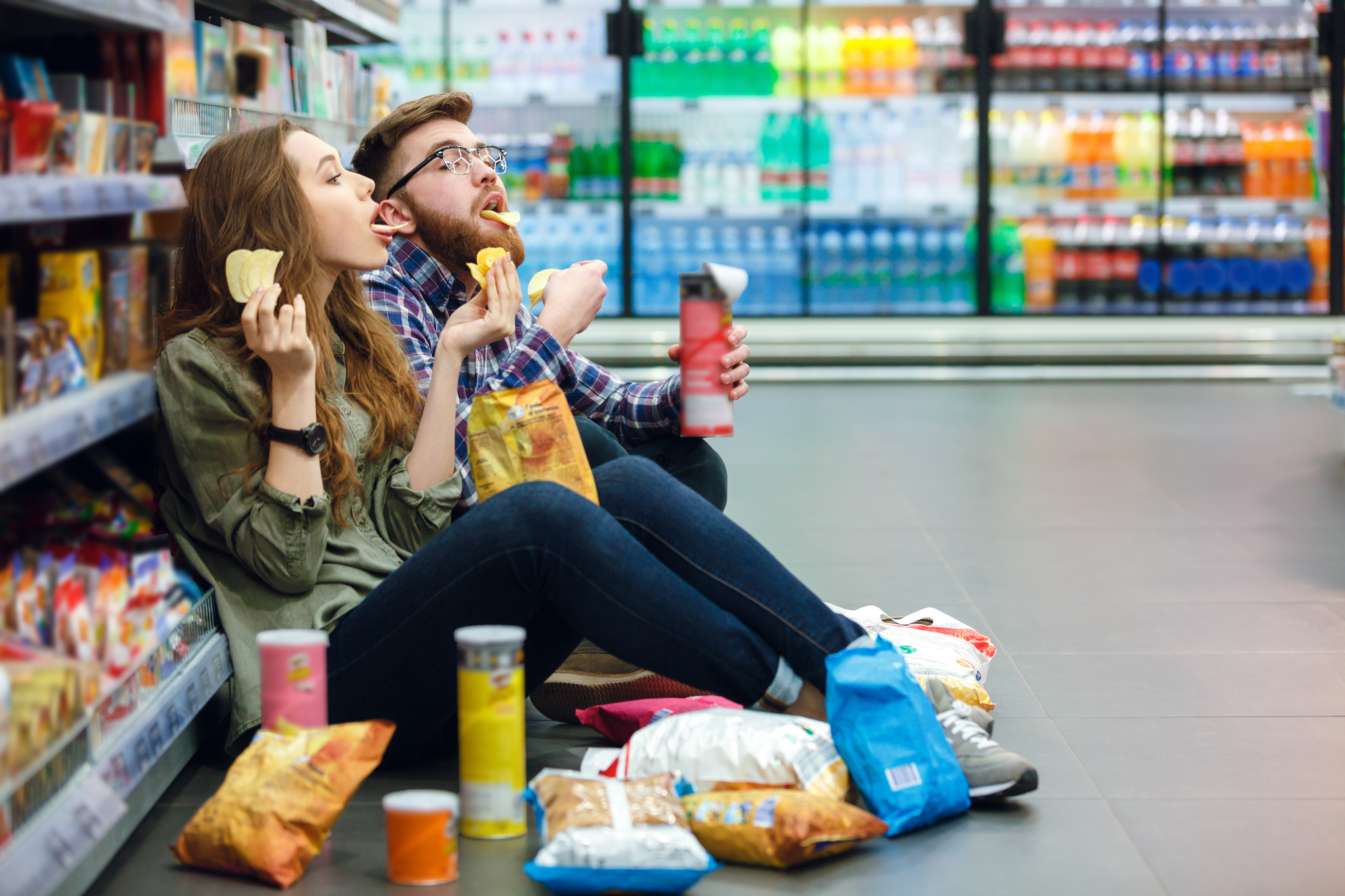 Giovane coppia seduta sul pavimento di un supermercato mangia snack e patatine; concept: junk food, cibo spazzatura