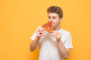 Young man holds a piece of fresh pizza in his hands to smell a scent with his eyes closed, isolated on a yellow background. Guy enjoys fast food on a yellow background.