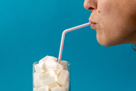 Profile of a young woman drinking with a red stripes straw from a glass filled with sugar cubes on blue background. Junk food, unhealthy diet, too much sugar on drinks, nutrition concept