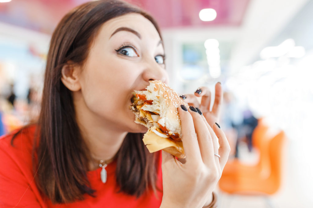 Pretty young funny woman eating hamburger inside in fast-food restaurant