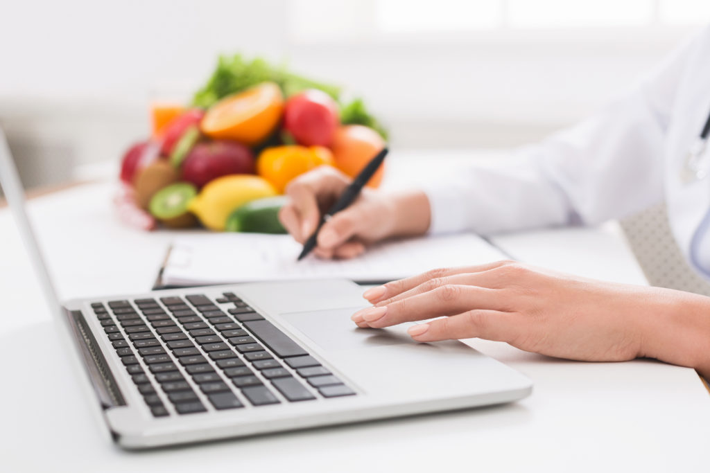 Nutrizionista, dietista o dietologa al computer mentre prende appunti su una cartella, sullo sfondo una pila di frutta e verdura; concept: dieta, nutrizionisti