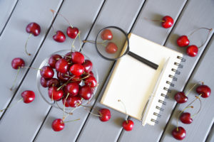 Ciotola di ciliegie con quaderno ad anelli, penna e lente di ingrandimento, e ciliegie sparse sul tavolo; concept pesticidi