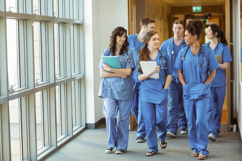 Six medical students or postgraduates in blue uniforms, notebooks and stethoscopes walk in a corridor with large windows