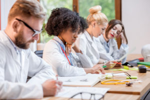 Gruppo multietnico di studenti di medicina in uniforme seduti insieme alla scrivania con diversi oggetti medici in classe