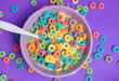 Cereali da colazione per bambini multicolorati e zuccherati in una tazza con latte e un cucchiaio; sul tavolo viola altri cereali sparsi