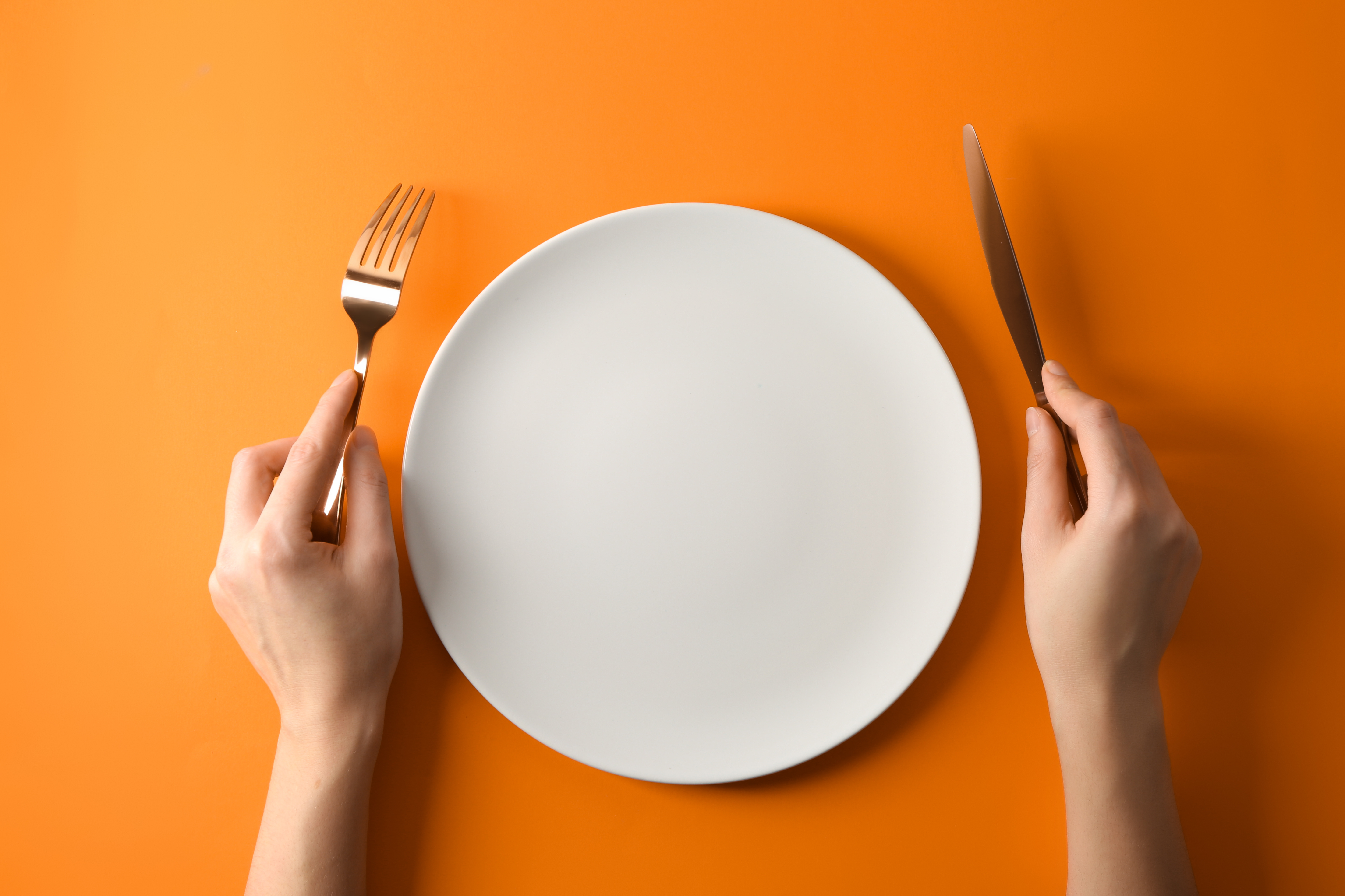 Mani femminili con coltello e forchetta attorno a un piatto vuoto; concept: digiuno, fame, dieta mima digiuno
