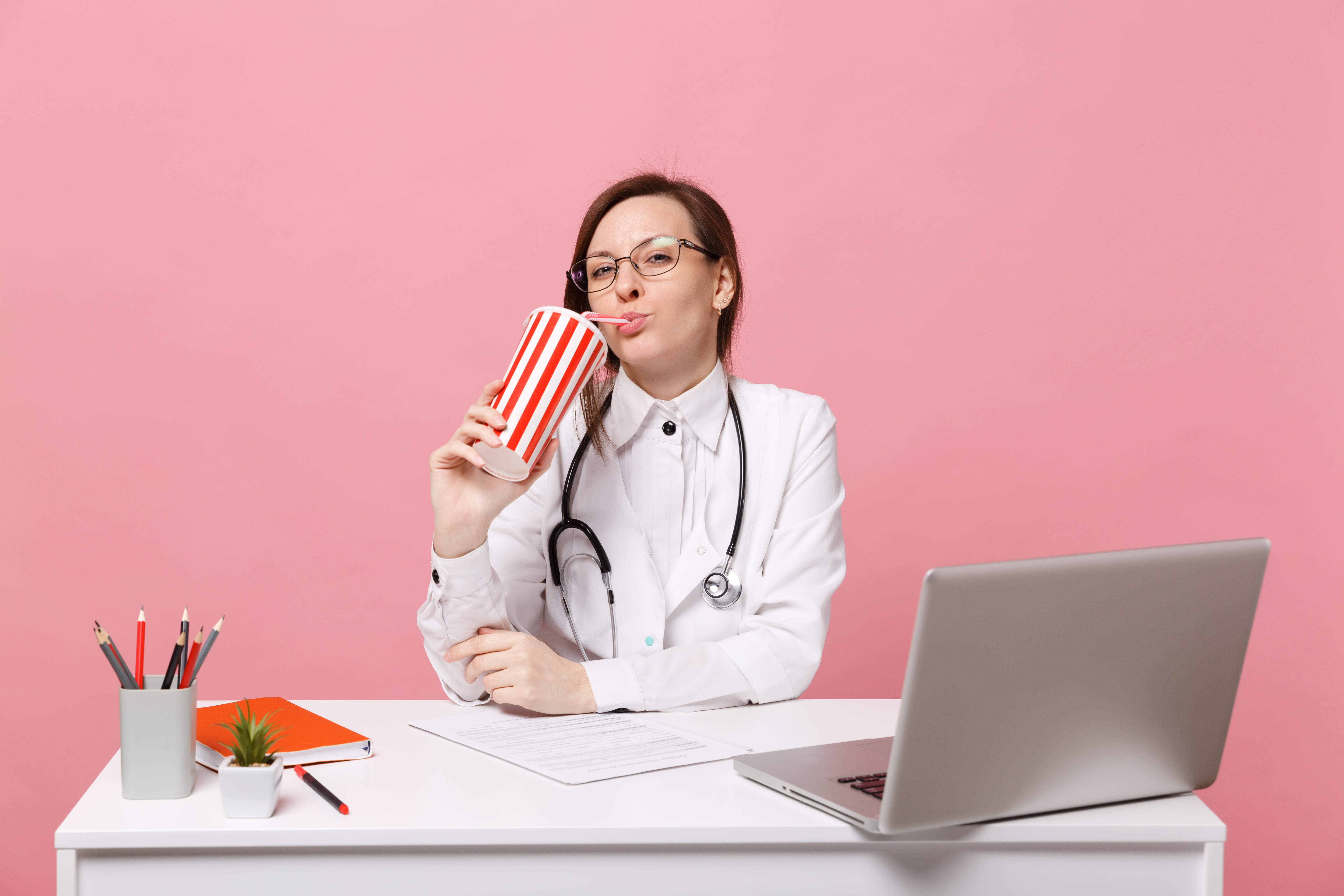 Female doctor sit at desk work on computer with medical document hold cola in hospital isolated on pastel pink wall background. Woman in medical gown glasses stethoscope. Healthcare medicine concept.