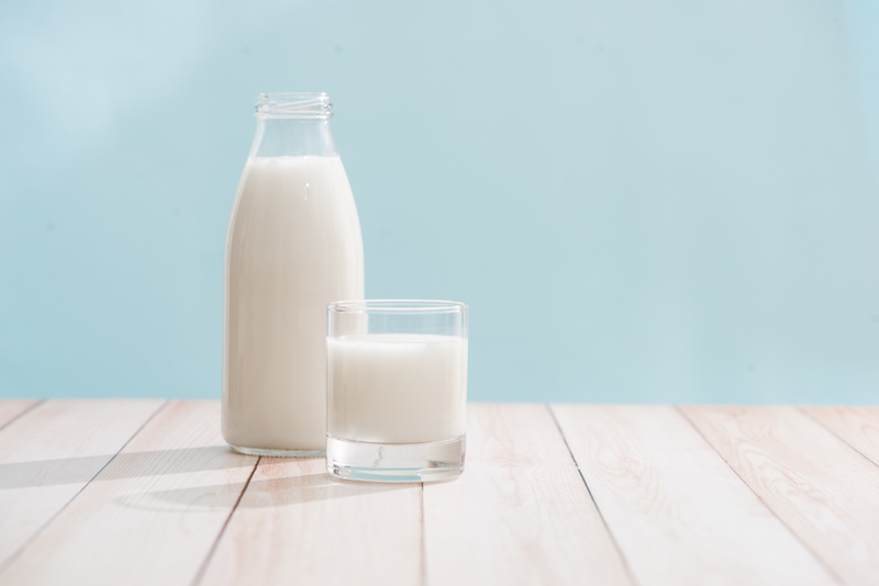 latte, bicchiere di vetro e bottiglia pieni di latte su piano di legno chiaro e sfondo azzurro