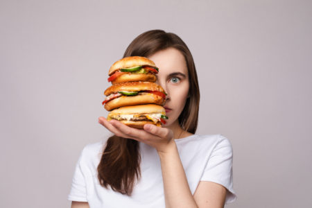 Una donna nasconde metà della sua faccia con tre panini con hamburger impilati su una mano; concept: alimenti ultra-trasformati junk food fast food cibo spazzatura hamburger