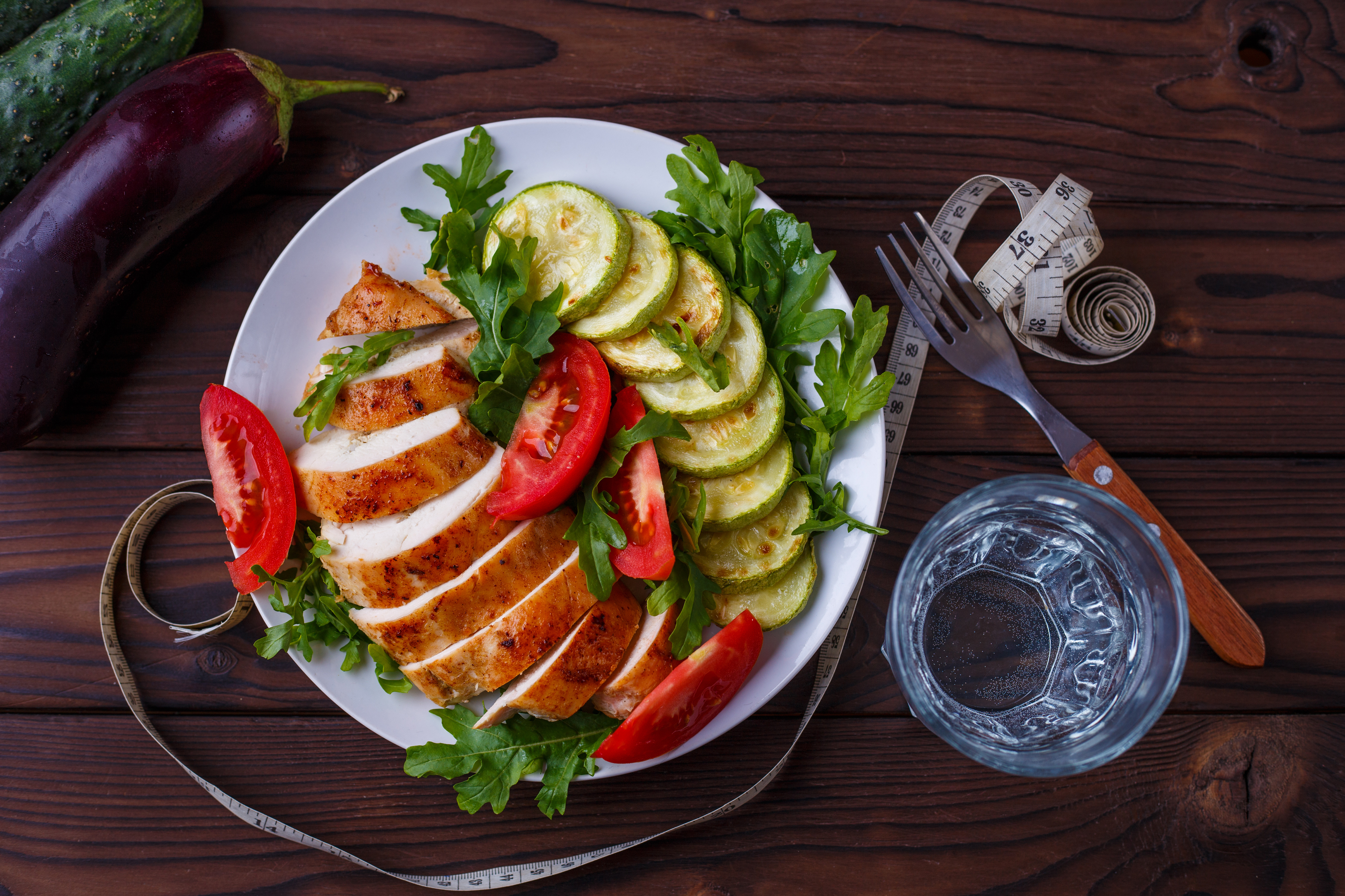 Piatto con pollo alla piastra, zucchine, rucola e pomodori, accanto a metro a nastro attorno a forchetta; concept: dieta