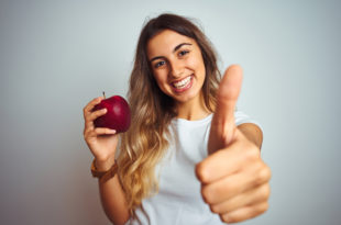 Young beautiful woman eating red apple over grey isolated background happy with big smile doing ok sign, thumb up with fingers, excellent sign