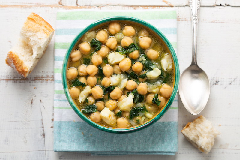 Ciotola di zuppa con ceci e verdure su una tovaglietta a righe bianche, verdi e azzurre, con un pezzo di pane a sinistra e a destra e un cucchiaio