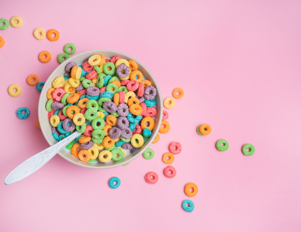 Cereali da colazione colorati in una tazza con cucchiaio, appoggiata su una superficie rosa; concept: zucchero, bambini