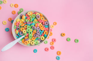 Cereali da colazione colorati in una tazza con un cucchiaio visti dall'alto; intorno altri cereali sparsi