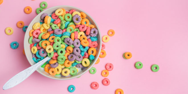 Cereali da colazione colorati in una tazza con cucchiaio, appoggiata su una superficie rosa