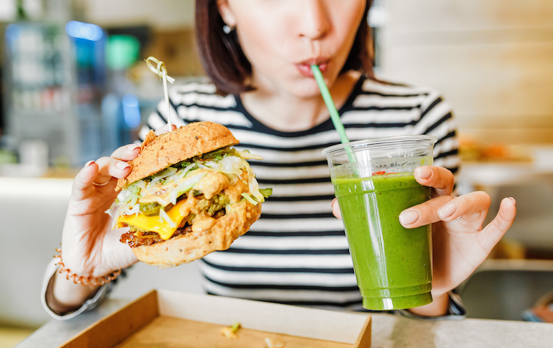 Ragazzo beve uno smoothie verde mentre tiene in mano un panino con burger vegetale