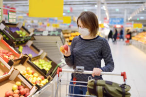 Donna con mascherina e carrello esamina una mela al supermercato; concept: ortofrutta, frutta