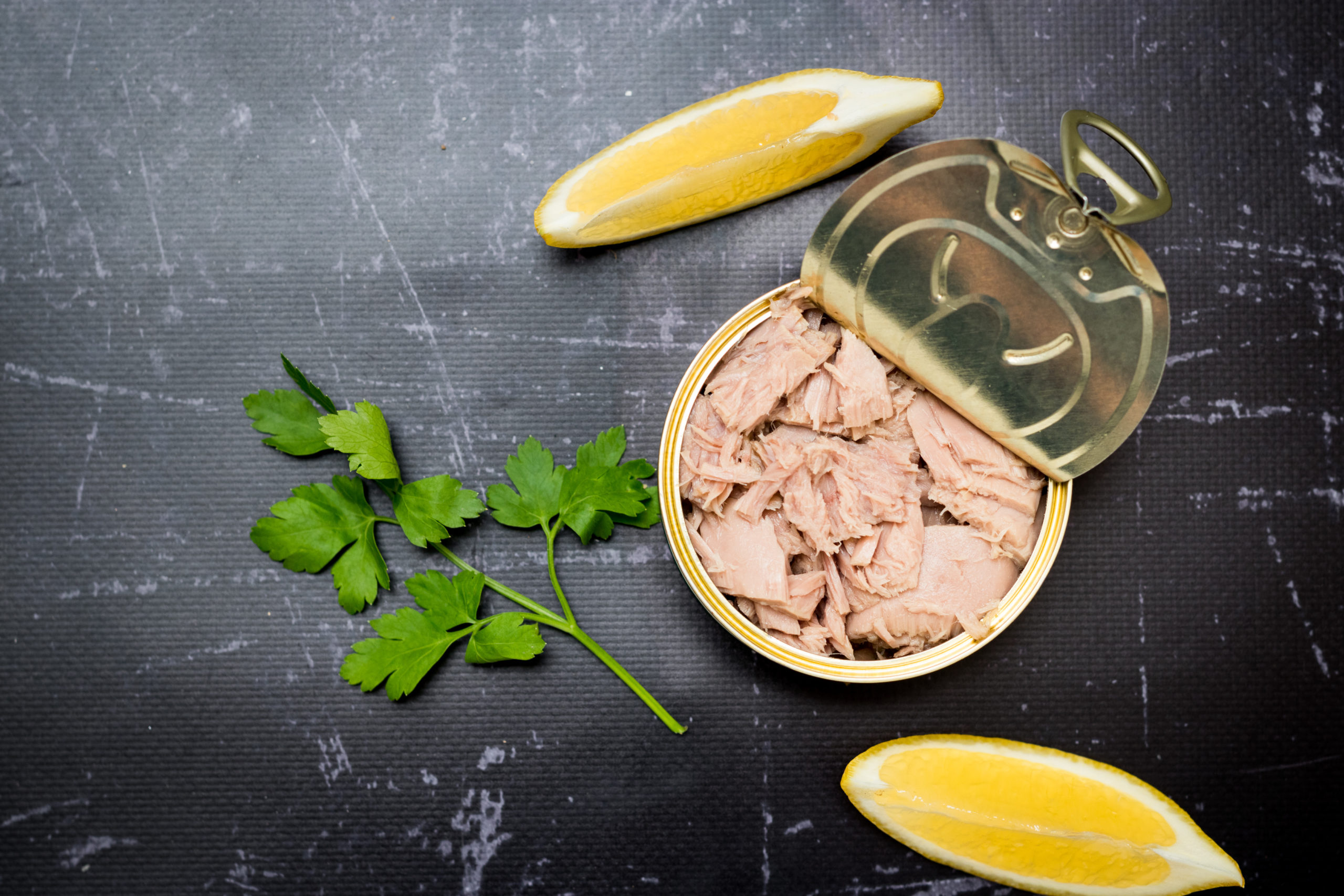 Canned tuna with lemon and parsley on dark background, top view.