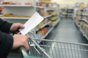 Una persona spinge un carrello tra gli scaffali del supermercato con la lista della spesa in una mano