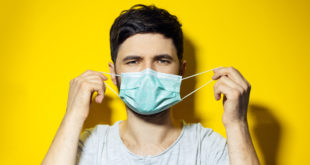 Studio portrait of young ill man, trying to put on, medical flu mask on yellow background mascherine