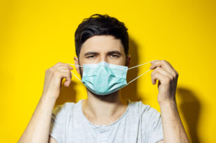 Studio portrait of young ill man, trying to put on, medical flu mask on yellow background mascherine