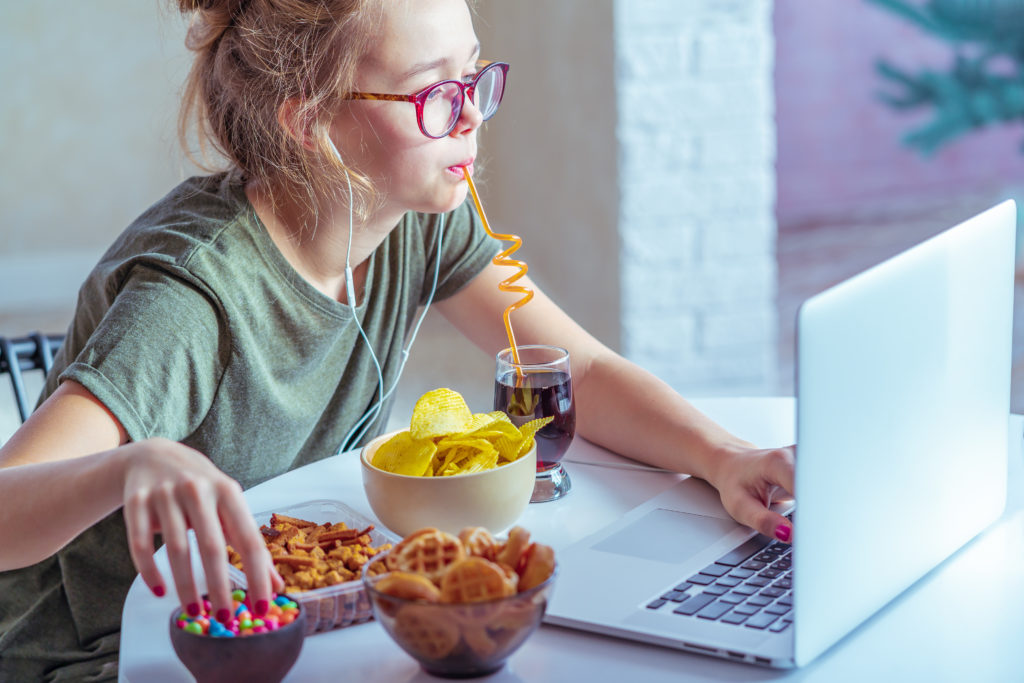 Girl works at a computer and eats fast food. Unhealthy food: chips, crackers, candy, waffles, cola. Junk food, concept.