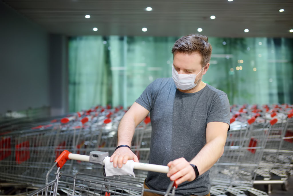 Man wearing disposable medical face mask wipes the shopping cart handle with a disinfecting cloth in supermarket. Safety during coronavirus outbreak.